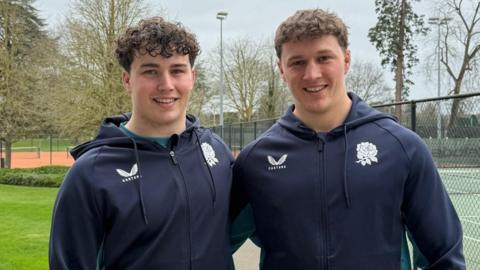 Brothers Declan and Connor Treacey smiling for a picture by a tennis court. They have their arms around each other.