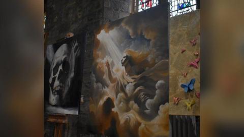 Three paintings depicting different artworks are displayed inside St Cuthbert's Church. There are stained windows in the background.