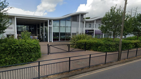 An exterior of the school taken from the road.  It is a new building with wave shaped roofs and large glass windows.