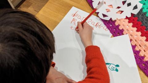 An over the head and shoulder view of a boy of about five years old, with short dark hair and wearing a red jumper writing to Father Christmas. In his right hand is a red pencil and he is writing in red on white paper, which is resting on a pine table. His top line says dear Santa