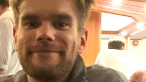 David Haw smiles at the camera in a small room, possibly a boat cabin. He has brown hair, cut short at the sides, and a beard.