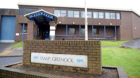 A general view of the entrace to HMP Greenock. A small, dark stone wall sits in front of the main building with the name of the prison on a white panel.