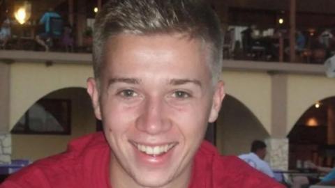 Sam Pegram smiles into the camera in a restaurant. Sam has short blonde hair and is wearing a red top.