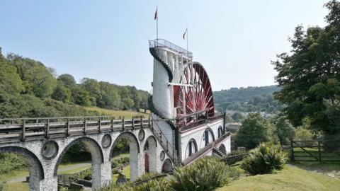 The wheel, which is red and has multiple spokes, sitting in its casing with viaducts leading to it carrying the rods that pump the water. It it surrounded by the green of the valley.