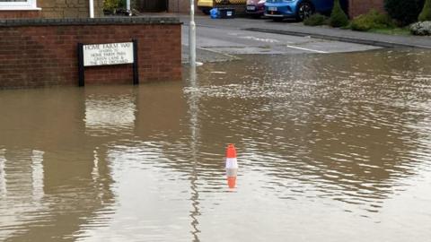 A view of a flooded suburban street. A street sign reads Home Farm Way. There is a cone sticking out from the middle of the water.
