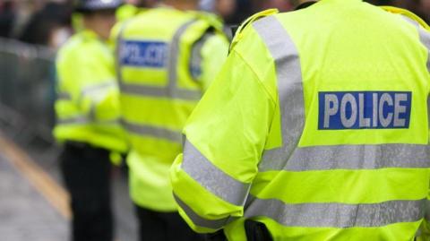 Three police officers standing outside wearing yellow fluorescent jackets with silver stripes and silver writing in a blue box which says POLICE.
