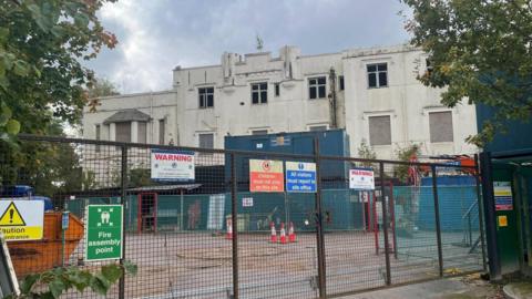 An exterior view of the three-storey white hotel, taken from behind a wire construction site fence with warning signs attached