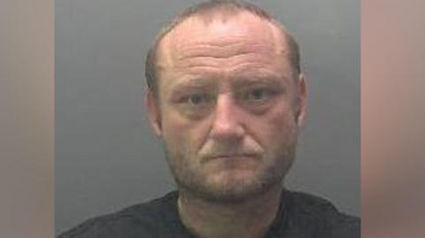 A custody image of Dean Wright, who has a beard, receding brown hair and is wearing a black top