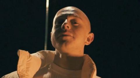 Young Spencer - a bald man is shot from below as he smiles at the camera. He is standing in the dark but an orange light is illuminating him. He is wearing a white puffer coat and a ribbed white t-shirt.