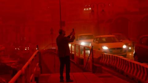 A man takes a picture while cars drive with their headlights on in red air in the city of Heraklion, Crete. Photo: 1 April 2026