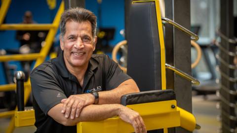 A man wearing a black polo shirt, is leaning on a yellow piece of gym equipment. He has short dark hair and smiles showing his teeth while his left hand rests on his right forearm.