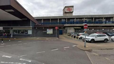 A wide grey and brown building with blue railings and a red national rail logo at the top. A number of cars and red no entry signs surround the building.