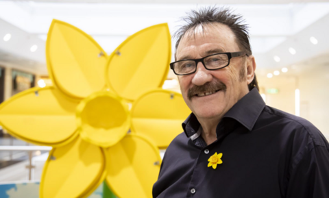 A man has black, short spiky hair, a moustache and black framed glasses. He is wearing a black shirt with a yellow daffodil pin. In the background is a large daffodil structure