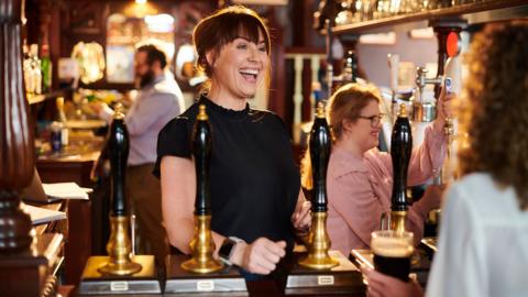 A woman smiling behind a car, as another woman pours a beer behind her.