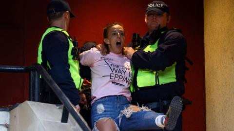 Sarah White is displaying an emotive expression as she is held by two police officers at the top of the stairs of a building. She is wearing a pink T-shirt that reads: "The Only Way is Epping". Two police officers in uniform stand either side of her and are touching her arms.