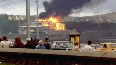 An archive photograph of the Summerland complex on fire, with thick black smoke billowing into the air. People look on from the promenade.