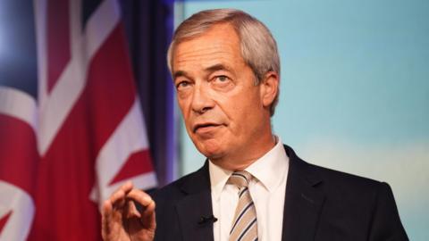 Reform UK leader Nigel Farage has grey hair parted to the side and is wearing a dark suit jacket, white shirt and striped tie