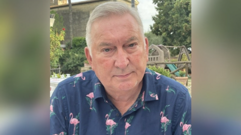 A head and shoulder image of a man - Dorian Jones - with grey hair wearing a navy blue skirt with pink flamingo on it looking down the camera lens with a white trees and a park in the background.