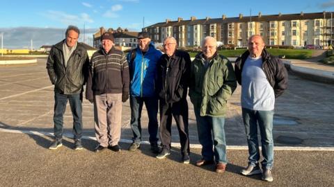 Six members of the boat club standing in the sunshine in front of the empty lake. 