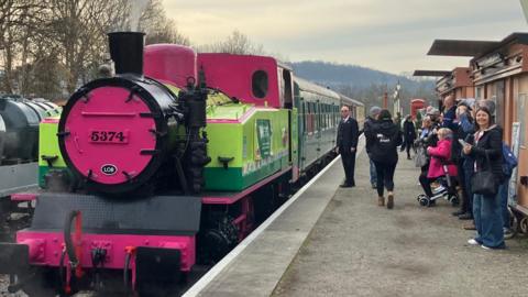 A picture of the steam locomotive about to set off at Plym Valley railway station. People can be seen looking at the train on the platform. The train itself is green in colour with hot pink at the front.