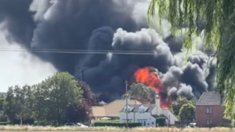 Large black smoke and orange flames are billowing into the sky from behind a building. In front of the building is a large open field with trees either side.