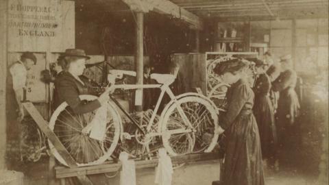 A black and white archive photo of women wearing long dresses and hats with their hair tied back as they hold a bicycle which is propped up on a stand. They are working on it. Behind them are other women working, while a man is on the far left.