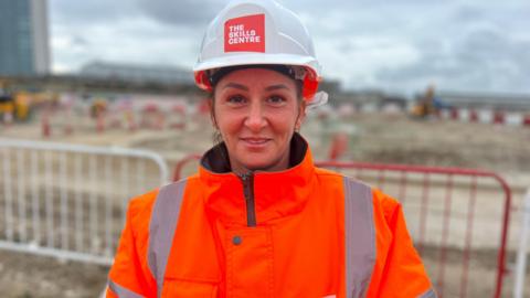 Charlie Lillington wearing an orange hi-vis jacket and hard hat with The Skills Centre written on it. A blurred building site and red and white metal fencing is behind her