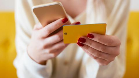 A stock picture of a woman holding a mobile phone and holding a yellow banking card.
