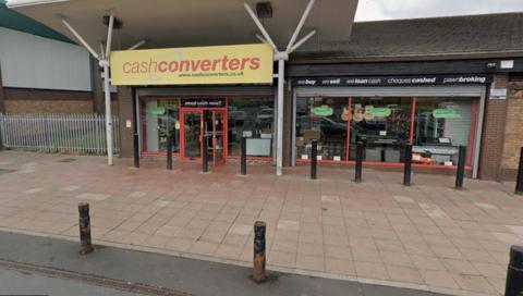 The front of a Cash Converters store. The sign indicating the shop is yellow, with red writing. A large pavement with bollards is visible in front of the store.