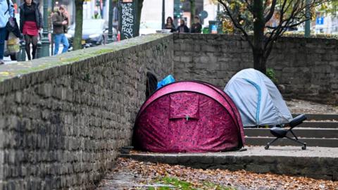 A pink and a blue tent are pitched on concrete steps. On the other side of the wall people walk past carrying shopping bags.