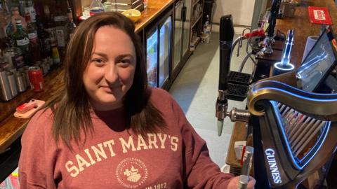 Megan stands behind the bar, smiling up at the camera. Behind her are bottles of alcohol and she stands next to the handles to pull pints. She has shoulder length brown hair and wears a salmon coloured hoodie with white writing on.