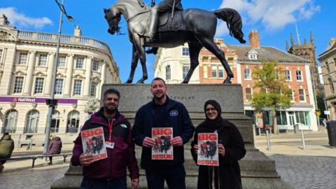 Two men and a woman stand in front of a statue, each of them holding a Big Issue magazine.