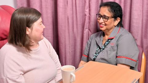 Nursing assistant Gurinder Cheema with a patient. Ms Cheema is wearing a grey and red uniform with a lanyard and is sitting at a table with a patient in a pink top, who has a cup of tea in front of her. The pair are smiling and chatting and there is curtain behind them.