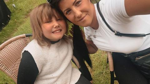 Summer and her mum Rebecca are sitting next to each other on wicker garden chairs. They're outside in the garden, Summer wears a black long sleeved t shirt with a white knitted sleeveless tank over the top, she has mousy blonde hair and smiles at the camera. Rebecca has black hair tied in a low side ponytail and has a stud in her upper lip. She wears a white short sleeved cotton t shirt and black leggings, with a black cross body leather bag.