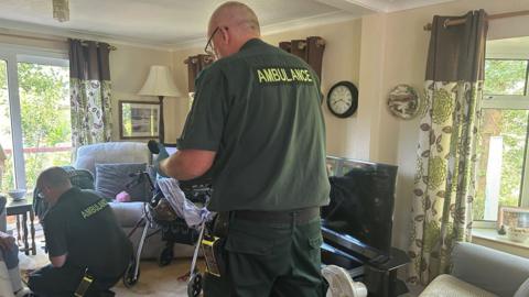 Two men in green South Western Ambulance Service uniforms treating a poorly woman in her front room.