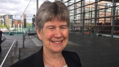 Jane Hutt photographed outside the Senedd, she has short silver hair, blue eyes and smiles towards the camera. She is wearing a black blazer and gold stud earrings and a gold slim chain necklace. 