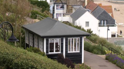 The pavilion - with black horizontal planks and white window frames and vertical supports - in the park with lavender plants and a clipped hedge outside. A footpath runs in front of the pavilion with houses and the beach seen in the background.