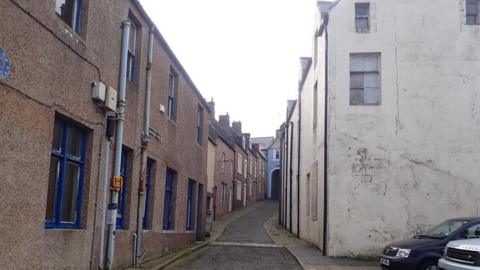A narrow lane with buildings up both sides and a cobbled pathway in between