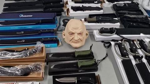 A table is filled with various knives and machetes sitting on top of their respective boxes. Some are wrapped in plastic packaging. In the centre of the image is a whole-head mask of a bald person.