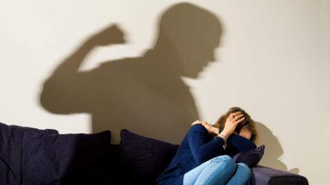A reconstruction has been choreographed for a photo depicting an example of domestic abuse. A woman is cowering on a sofa protecting her head. The shadow of a man is cast on the wall, appearing to raise his fist ready to punch.