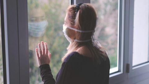 A woman wearing a surgical mask stands looking out of a window. She has her left hand placed on the glass, and is wearing a black top. Her long hair is held back by a headband.