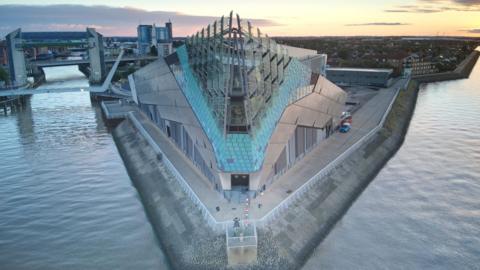 A striking modern building in a triangular shape, with a glass front above blue and silver tiles, juts out on a promontory between two rivers under a yellow sky. To the left, bridges and a H-shaped tidal barrier can be seen straddling a river. To the right, houses can be seen stretching towards the horizon. 