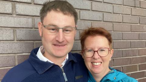 Richard Foggin and Tracy Hanlon from Swindon smiling both wearing glasses stood in front of a brown brick wall at the current bus station in Swindon.