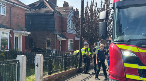 The photo shows a street of houses. The side of one of the buildings has been scorched black. There are also holes in the side of the roof. There are three police officers in hi-vis jackets, and a fire officer in a blue polo top. There is also a fire engine in the forefront of the photo.