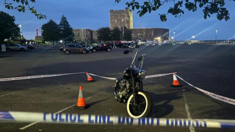 A black Harley-Davidson motorbike at a car park sectioned off with cones and police tape. It is night, a castle can be seen in the background and some street lights.