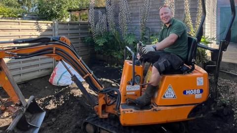 A man sat on a digger which is orange with the bucket of the digger resting on the floor. The man wears black shorts and a green polo shirt. The digger is situated in a back garden