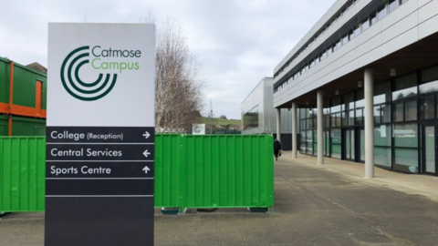 A school building with a large sign saying Catmose College, and a logo, in the foreground with a green metal fence just behind it 