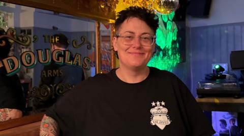 A woman with short black hair and glasses stands in front of a mirror with "The Douglas Arms" written across it in gold