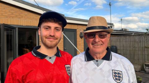 Jacob (the grandson) is on the left, it's a sunny day and he's in a blue England cap with a red England shirt on. He has brown hair and blue eyes, he's smiling. To his left is his grandad Geoff he has sunglasses on and a thatch hat. He looks happy and is in a matching England shirt which is white. 