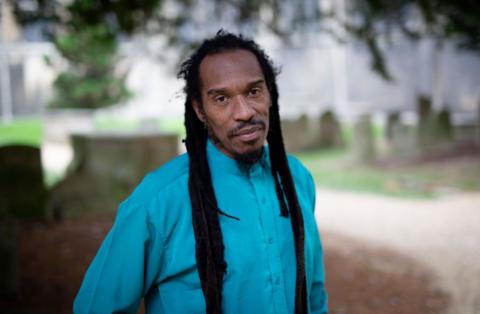 Benjamin has long dark brown dreadlocks and is looking into the camera with a small smile. He is wearing a bright blue shirt.